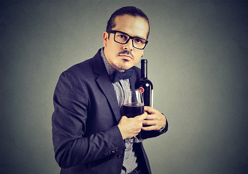 Man in suit and glasses holding bottle and wineglass with feeling of ownership and looking at camera.