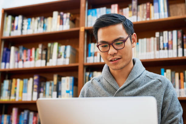 He only strives for success through hard work Shot of a focused young man busy working on his laptop and studying while being seated inside of a library asian adult student stock pictures, royalty-free photos & images