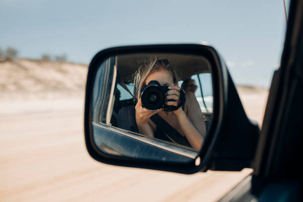 Young woman taking a selfie with a DSLR in the wing mirror of a 4x4 while driving along the 75 Mile Beach on Fraser Island, Australia.