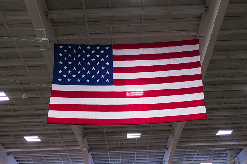 An large american flag hanging from the rafters of an indoor arena.