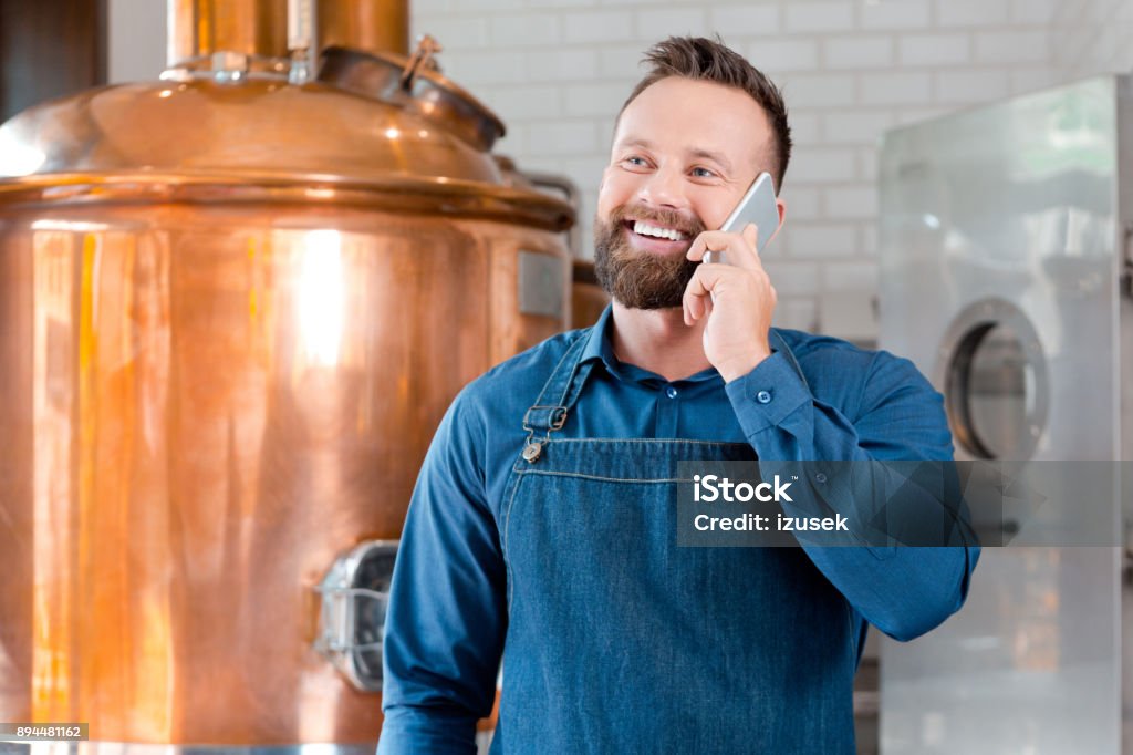Master brewer talking on mobile phone in brewery Master brewer talking on mobile phone in fermenting section of beer manufacturing factory. Mature man talking on phone in his microbrewery Owner Stock Photo