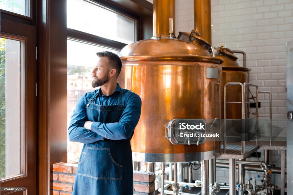 Master brewer standing in micro brewery Master brewer standing with his arms crossed and looking away in micro brewery. Mature man in uniform and apron standing in beer fermentation section. Brewery Stock Photo