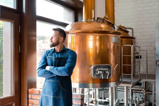 Master brewer standing with his arms crossed and looking away in micro brewery. Mature man in uniform and apron standing in beer fermentation section.