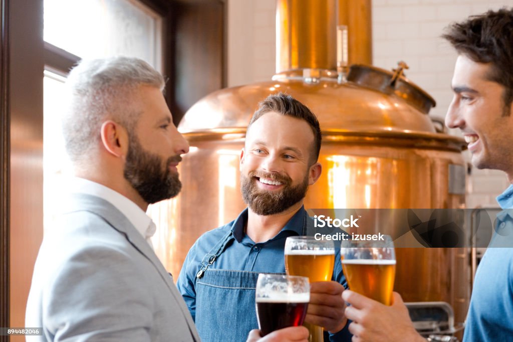 Smiling men toasting beers at micro brewery. Smiling men toasting beers at micro brewery. Team of three people toasting beer glasses at brewery. Celebratory Toast Stock Photo