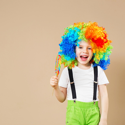 Happy clown boy in large colorful wig. Let's party! Funny kid clown. 1 April Fool's day concept. Portrait of a child eating lollipop. Birthday boy.