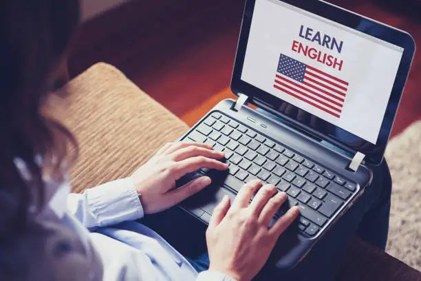 Woman sitting at home with a laptop on the knees and a website to learn American English on the screen.