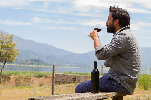 Latin young good looking handsome backpacker traveler male with beard, drinking in a fine glass cup with red wine served by generic wine glass bottle, in a lake shore in Chilean Patagonia, wearing a grey jacket and blue jeans. On back, Los Andes mountain range near to Torres del Paine National Park and Bariloche, Argentina in a sunset afternoon. Medium Shoot landscape with a copy space.