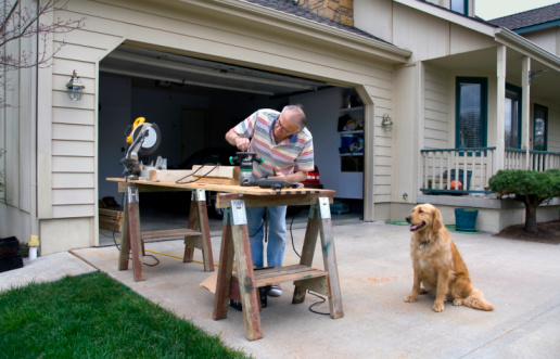 A retired craftsman adjusts a woodworking router on a temporary workbench in his suburban Missouri driveway as his Golden Retriever sits nearby.