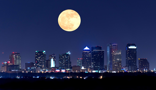 Night view of the Tampa Florida skyline showing skyscrapers with lights and a huge full moon in the sky over the buildings.