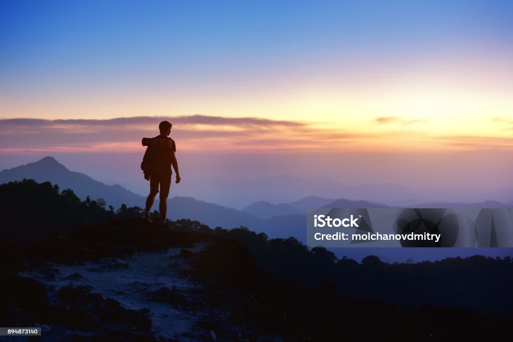 Man's silhouette on sunset mountains backdrop Silhouette of man with backpack at mountain top on background of sunset mountains Mountain Stock Photo