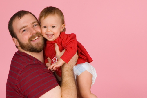 Proud father holds a smiling baby girl for a family portrait against a pink background.