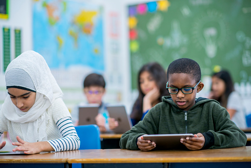 A Muslim girl and a boy of African descent are sitting beside each other in a classroom. They are working with tablet computer. They are learning about technology.
