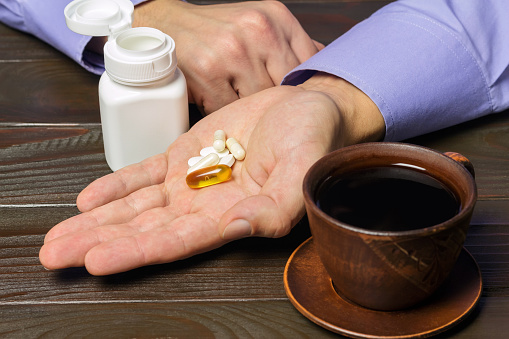 Male hand holds vitamin complex and dietary supplement, capsules of omega 3, glucosamine, calcium pills, cup of black coffee on dark wooden table.