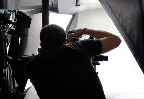 View from behind of photographer at work in his studio. He has a camera mounted on a tripod and is seen looking through the viewfinder.  Various lights are also seen to his left and right.