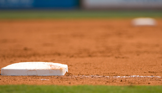 This photo is a close up of third base on a Baseball field at a major league or little league baseball game. the third base is white and there is the infield dirt and second base in the background. this is at a live sporting event. and this is an abstract background. 