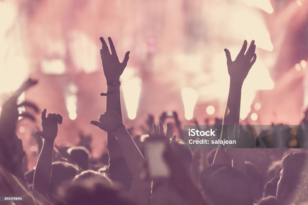 Crowd of audience with hands raised at a music festival Crowd of audience with hands raised at a music festival. Lights streaming down from above the stage Applauding Stock Photo