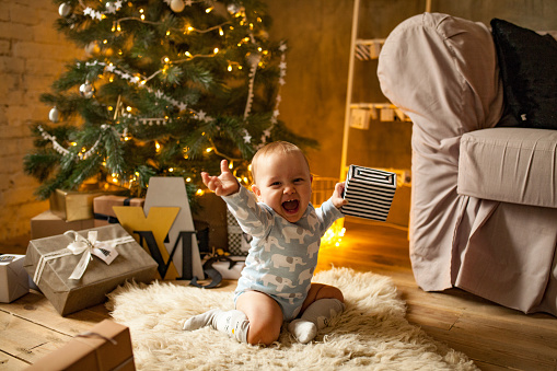 Little baby boy is happy with Christmas gift. He sits, laughs cheerfully and holds Christmas gift in his hand.