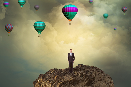 Businessman on cliff looking up at flying hot air balloons