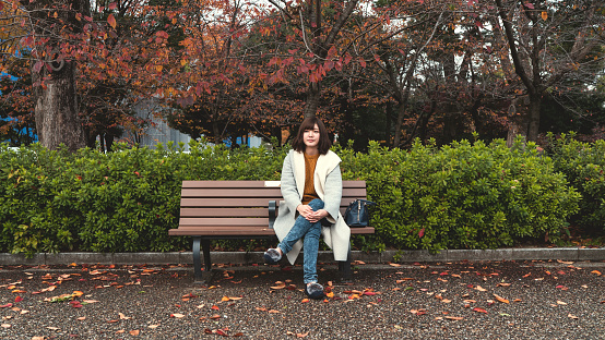 There is a young woman sitting on a bench at a park. There are many leaves on the ground and trees are turned red in autumn.