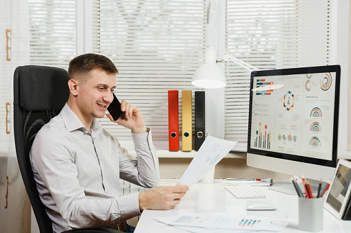 Smiling business man in shirt sitting at the desk, talking on mobile phone, conducting pleasant conversation, working at computer with modern monitor, documents in light office on window background