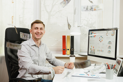 Handsome smiling business man in shirt sitting at the desk, throwing lot of cash money, working at computer with modern monitor, documents in light office on window background. Manager or worker