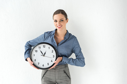 Smiling businesswoman holding wall clock in hands on white