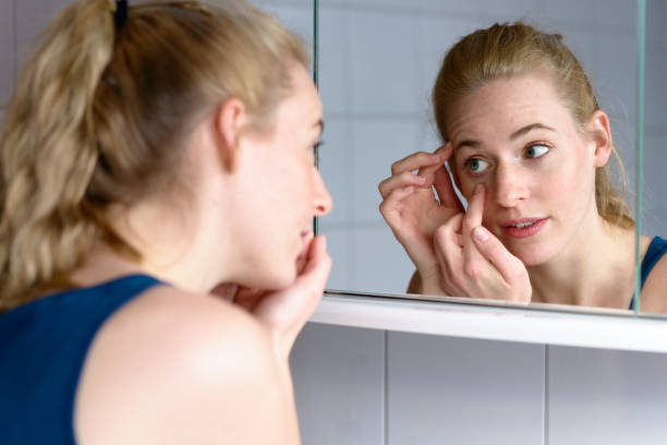 Young woman worried while looking in the mirror Over the shoulder view of a beautiful young woman worried about aging while looking in the mirror at the skin around her eyes lens eye stock pictures, royalty-free photos & images