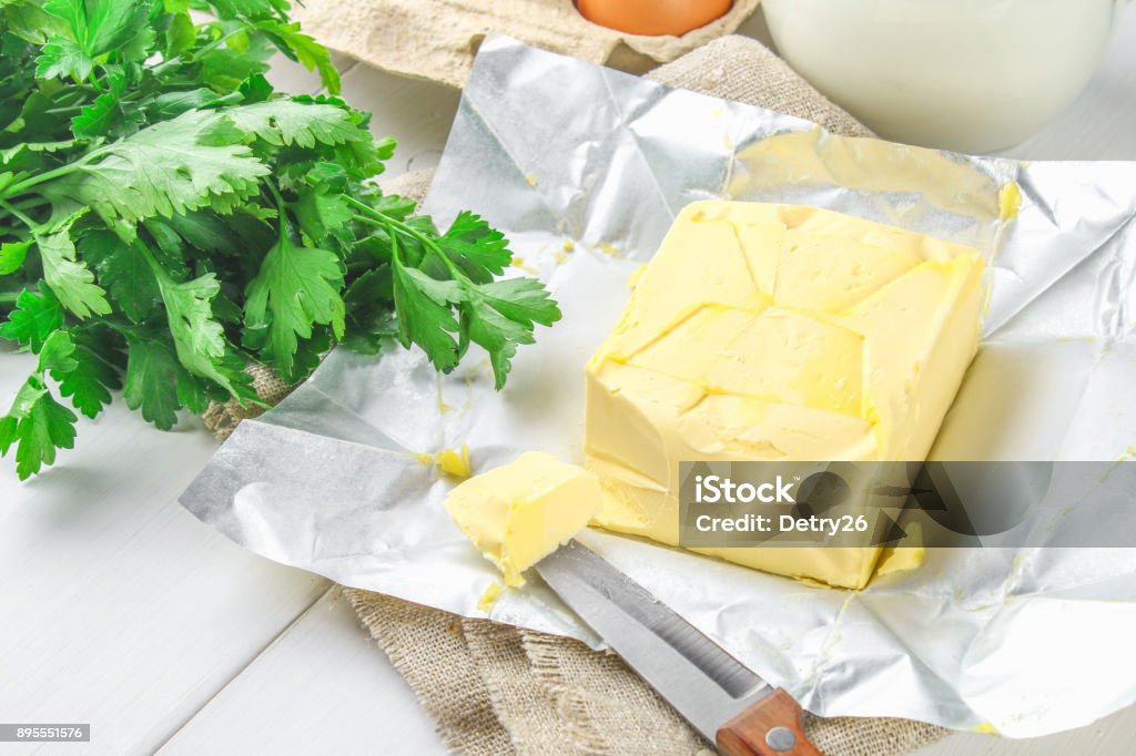A bar of butter is cut into pieces on a wooden board with a knife, surrounded by milk, eggs and parsley on a white table. Ingredients for cooking. A bar of butter is cut into pieces on a wooden board with a knife, surrounded by milk, eggs and parsley on a white table. Ingredients for cooking Basil Stock Photo