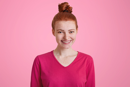 Happy smiling frecked female with red hair tied in knot, being glad to recieve compliment, isolated over pink background. Beautiful woman wears casaul clothes going to have walk outdoor with friends