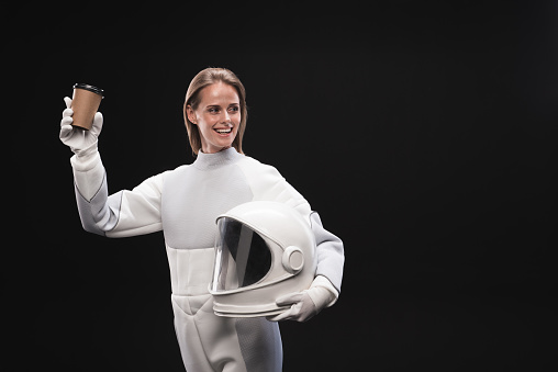 Best beverage. Cheerful young charming spacewoman wearing protective suit is enjoying hot coffee. She is looking aside with wide smile while standing and holding helmet. Isolated with copy space