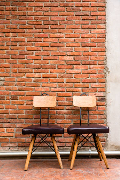 Two steel frame chairs are padded with brown cushions and a wooden backrest Two steel frame chairs are padded with brown cushions and a wooden backrest is placed in front of the brick wall backrest stock pictures, royalty-free photos & images