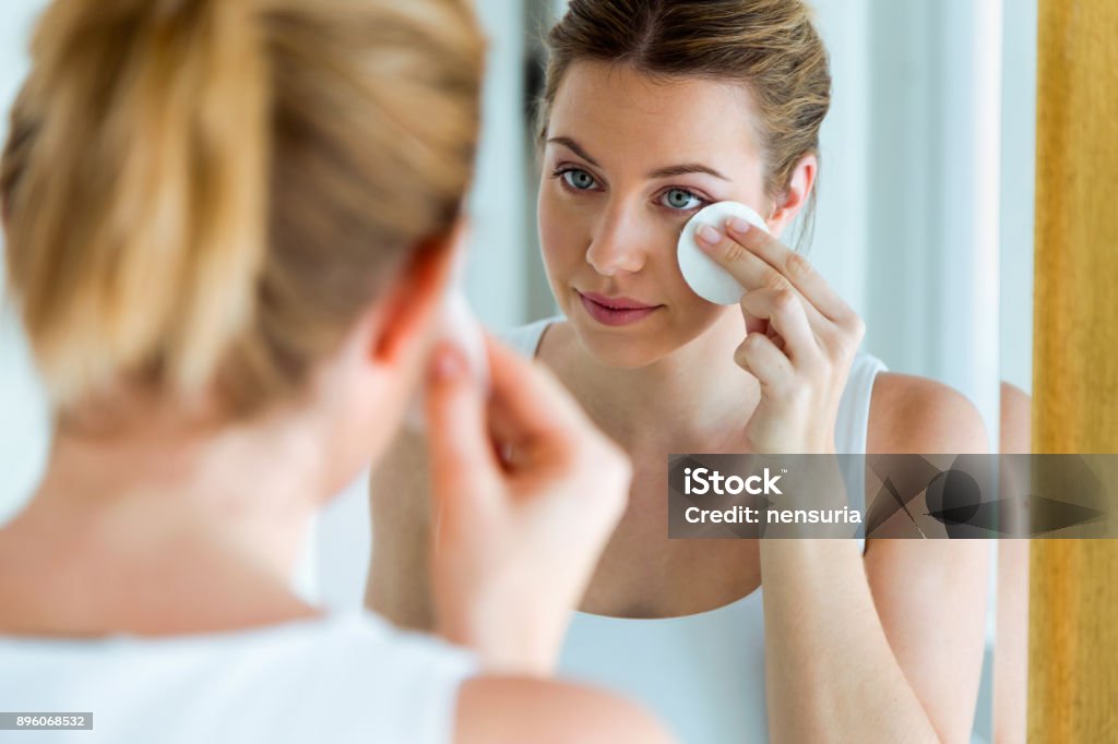 Hermosa joven está limpiando su cara mientras se mira en el espejo en el baño. - Foto de stock de Quitarse el maquillaje libre de derechos