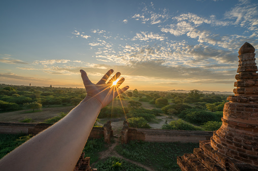 Pov of human hand stretching towards the sun at sunrise. Sunbeam effect, Bangan ancient temples on background. Energy people environment concept.