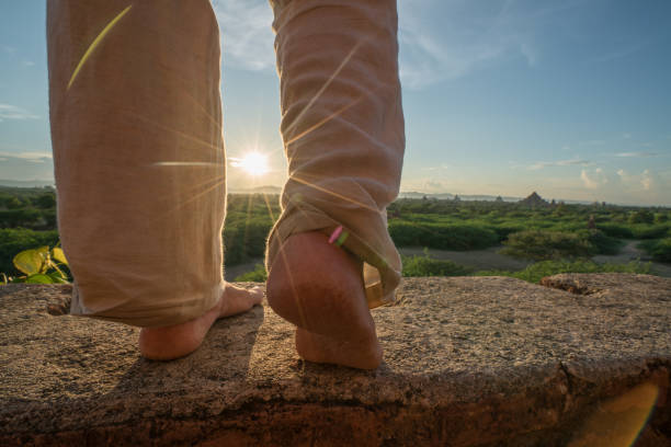 Young woman walking on temple's roof in the Bagan archeological zone at sunrise, close up on barefoot, temples pagodas and monastery coming out from the lush greenery. People travel concept.