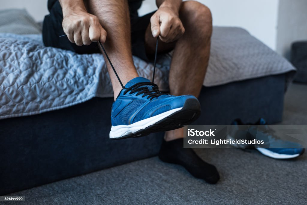 man tying sneaker laces Cropped shot of man sitting on bed and tying laces on jogging sneakers Men Stock Photo