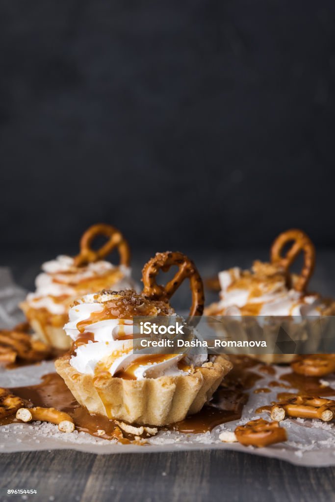Homemade cupcakes with salted caramel and pretzels. Sweet cakes with syrup and cream cheese on baking paper and wooden table. Baked Stock Photo