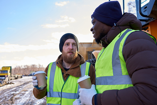 Portrait of two workers, one African-American, drinking coffee and chatting next to heavy industrial truck on worksite, copy space