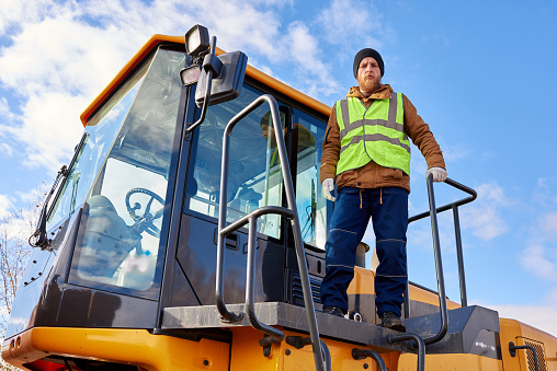 Low angle portrait of bearded worker standing on heavy yellow truck posing looking at camera against cold blue sky