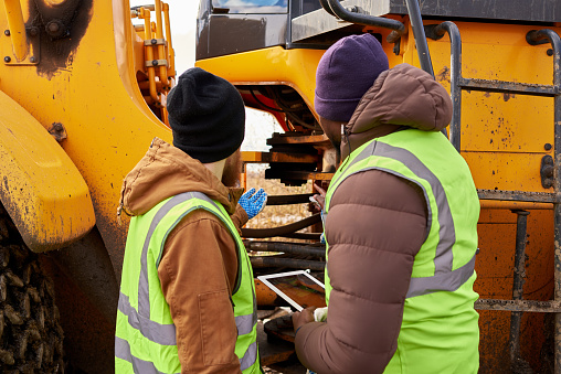 Back view portrait of two workers wearing reflective jackets, one of them African-American, discussing job on industrial site outdoors repairing big yellow truck