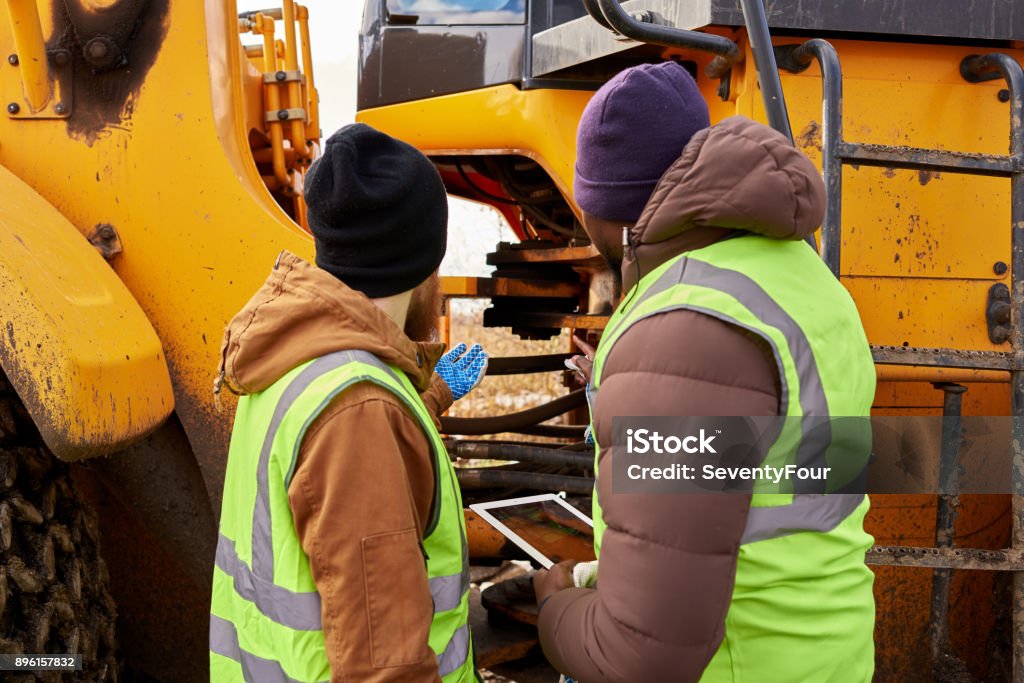 Mechanics Inspecting Truck Outdoors Back view portrait of two workers wearing reflective jackets, one of them African-American, discussing job on industrial site outdoors repairing big yellow truck Repairing Stock Photo