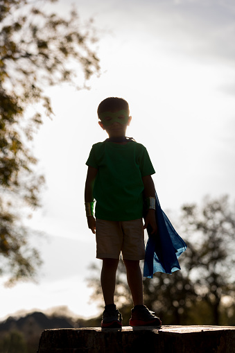 In this back lit photo, a little boy stands on a platform in the sunlight outside wearing  shorts, a t-shirt and a mask and cape.