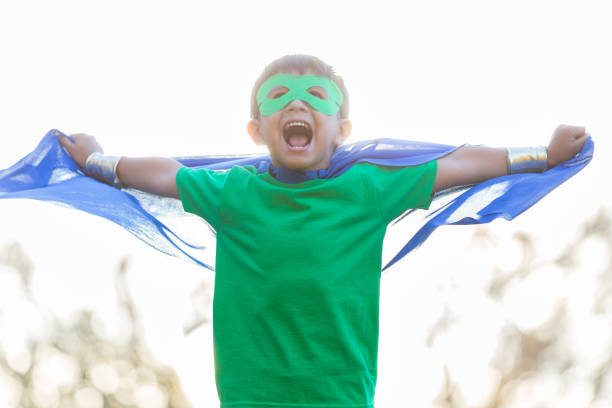 In this low angle view, a little boy is playing superhero outside.  He wears a mask and cape as he spreads his cape with outstretched arms and yells his battle cry.