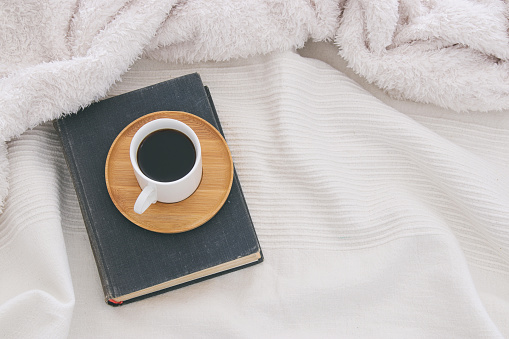Cup of coffee on the old book over cozy and white blanket. Top view