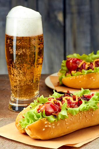 Two hot dogs with bacon and onion topings, glass of beer on stone table