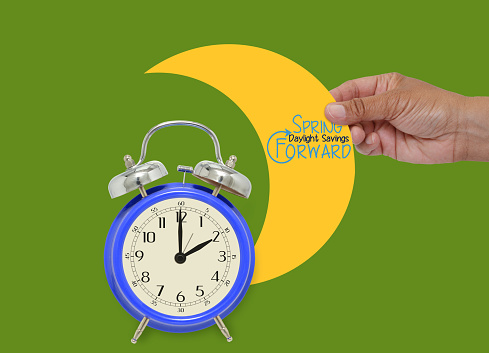2 o'clock alarm blue in front of half moon Spring Forward Daylight Savings Time held in fingers on green background