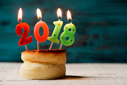 closeup of four lit number-shaped candles of different colors forming the number 2018, as the new year, on a cheesecake on a table, with a blank space on the right