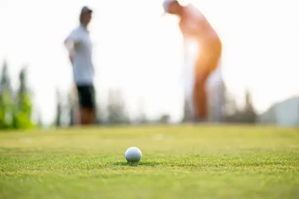 Photo of Golf ball approach to the hold on the green.  Couple golf player putting golf ball in the background.  Lifestyle Concept.