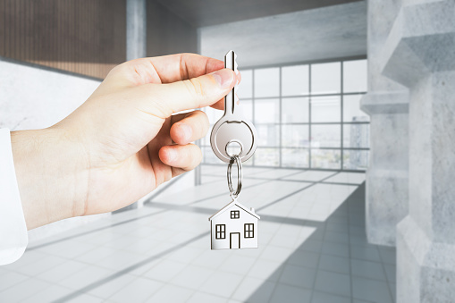 Businessman hand holding key with house keychain on abstract concrete interior background. Office and Real Estate Agent concept. 3D Rendering