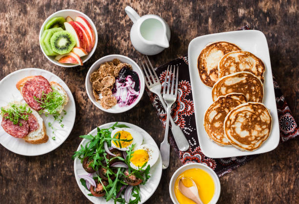 Breakfast table - greek yogurt with whole grain cereals and berry sauce, pancakes, arugula, cherry tomatoes, boiled eggs salad, kiwi, apples fruit, salami and cream cheese sandwiches on a wooden background, top view. Flat lay desayuno mesa - yogur griego con cereales de grano entero y salsa de bayas, panqueques, rúcula, tomates cherry, cocido, ensalada de huevos, kiwi, fruta manzanas, salami y sándwiches de queso crema sobre un fondo de madera, vista superior. endecha plana