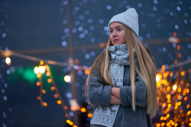 Contemplative young woman in hat and scarf with deer keeping hands in sweater sleeves while standing alone at Christmas night